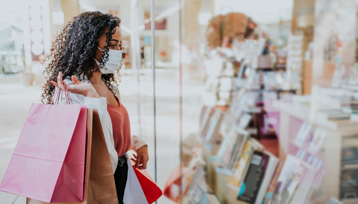 young woman with black curly hair wearing a mask and holding retail bags looking at a retail window