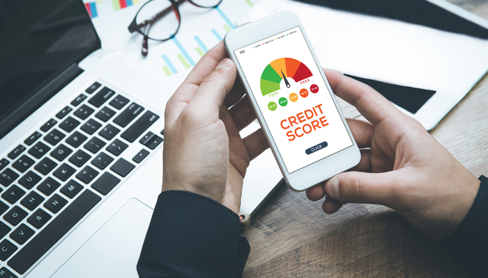 hands leaning on desk holding mobile phone with credit score rating on it and laptop, glasses and paper on desk