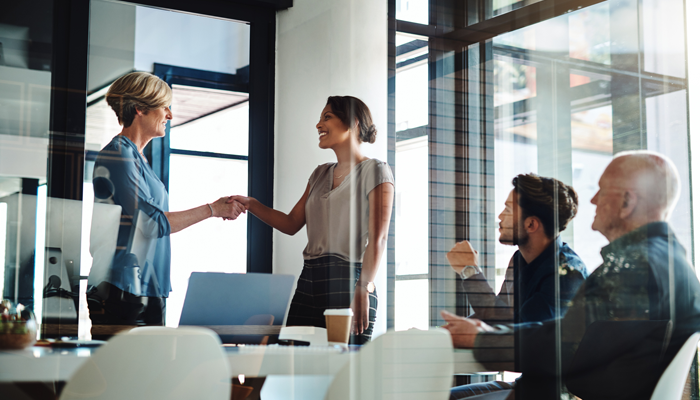 2 men sitting at office desk while 2 women standing in the same office shaking hands indicating recruiting post pandemic