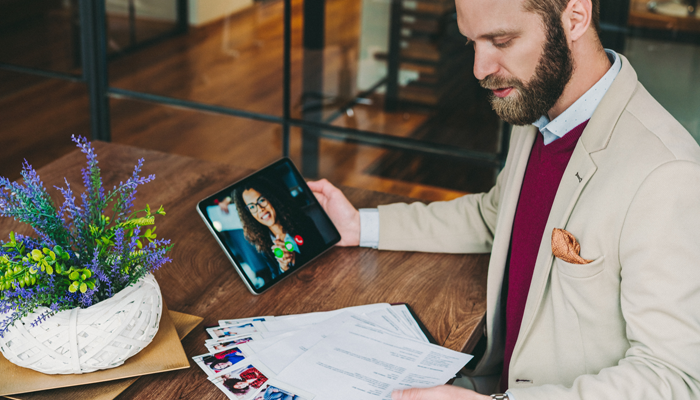 well dressed man holding tablet with an woman on a zoom and a bunch of resumes in scattered on desk and in his hand indicating recruiting post pandemic