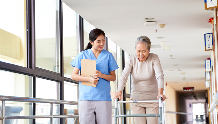 young asian nurse holding clipboard walking alongside an elderly woman with a walker