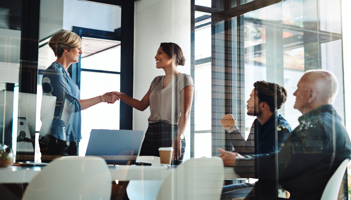 2 men sitting at desk and 2 standing women shaking hands in an office looking like one is prepared to enter job market
