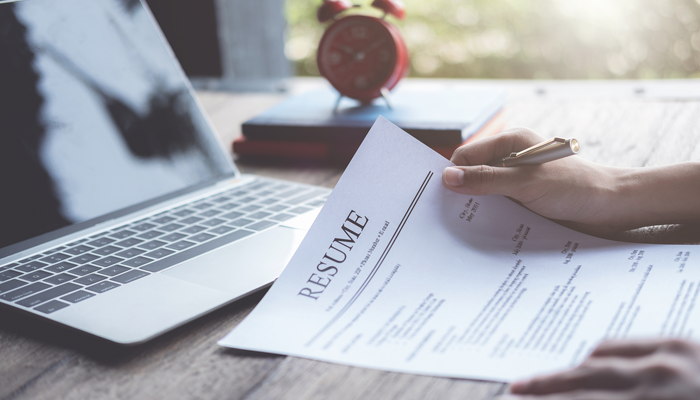 female hands on desk holding pen and paper resume with laptop and alarm clock on stacked books on desk