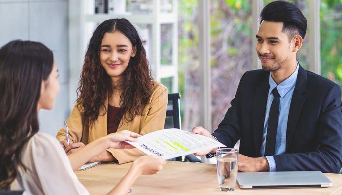 3 young professionals sitting around a desk and one male handing over his resume after completing a education history verification