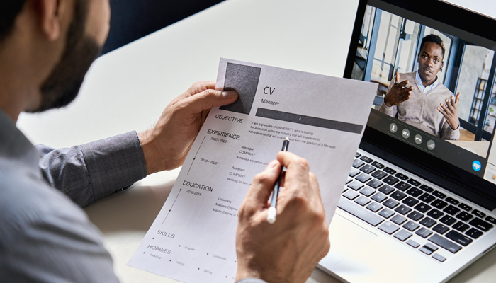 black man on laptop screen being interviewed and a man sitting at desk interviewing him holding a CV completing a reference check