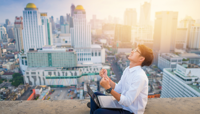 young asian man sitting overlooking the city with laptop on his lap happy and holding fists up in excitement for the best place to find a job in canada