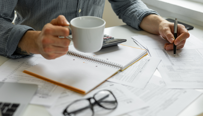 close up of a desk filled with papers calculator glasses and man hands holding a coffee and a pen in other hand looking over financial management jobs