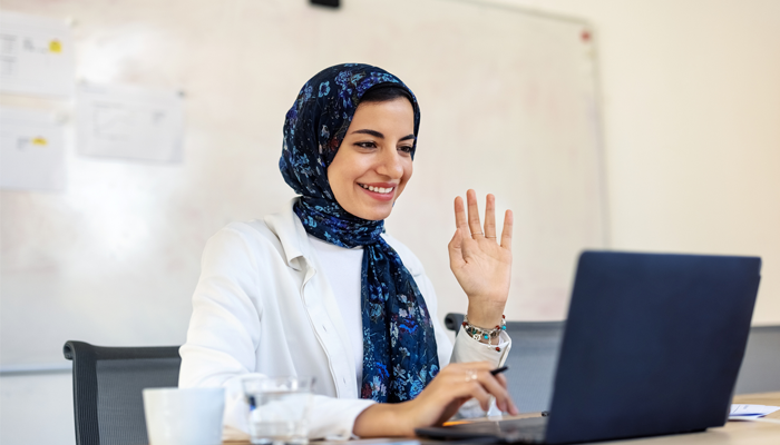 young woman wearing hijab waving to another professional on laptop screen showing hybrid work model