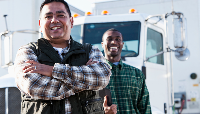 2 happy male truck drivers at their truck driving jobs with trucks in the background, one with hands crossed and one holding a clipboard