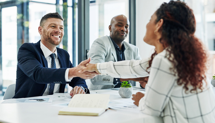 professional woman sitting across a desk from 2 professional men shaking the hand of one of them in job interview format for the ideal employment
