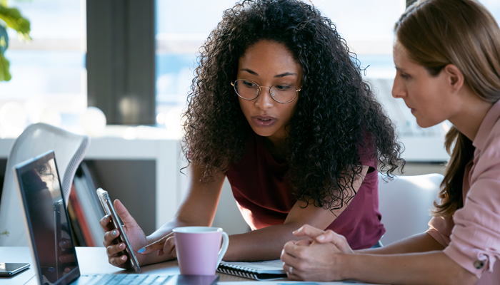 2 young women sitting at desk looking and pointing at calculator discussing pay transparency