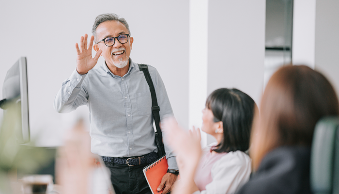 elder asian man carrying bag and notebook waving at a team of coworkers showcasing seniors in the workplace