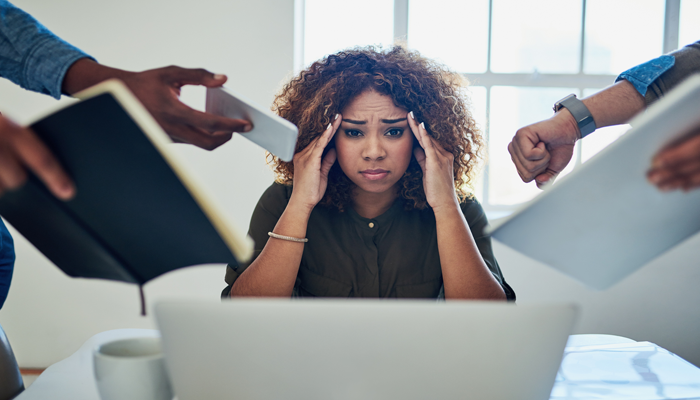executive burnout woman with overwhelmed look on her face sitting at desk with computer in front of her, hands to her temple and a bunch of hands reaching out to her with things to oversee