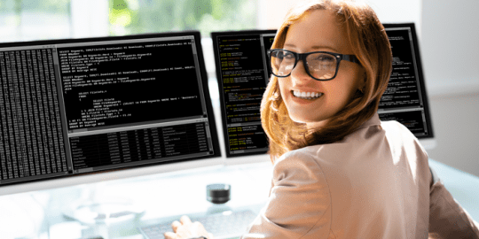 woman with a software engineer job in canada looking over shoulder smiling at camera from 2 big monitor screens with data on them