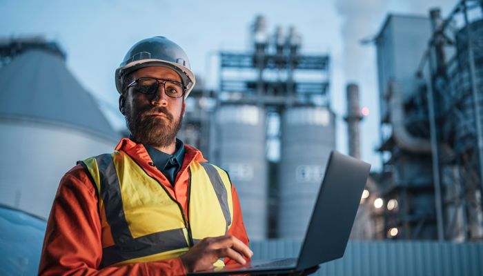 energy and utilities worker with hard hat, construction vest and glasses holding a laptop with utility structures in background