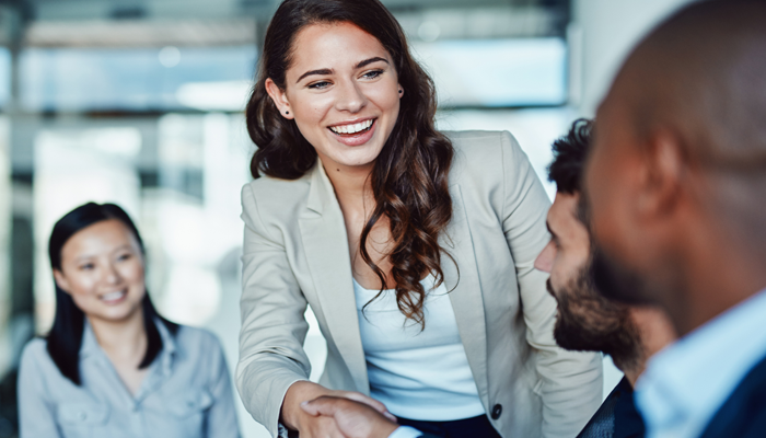 professionals sitting around a desk and an in focus woman shaking the hands of one of the executives. seems like she was just hired by the recruitment and staffing team