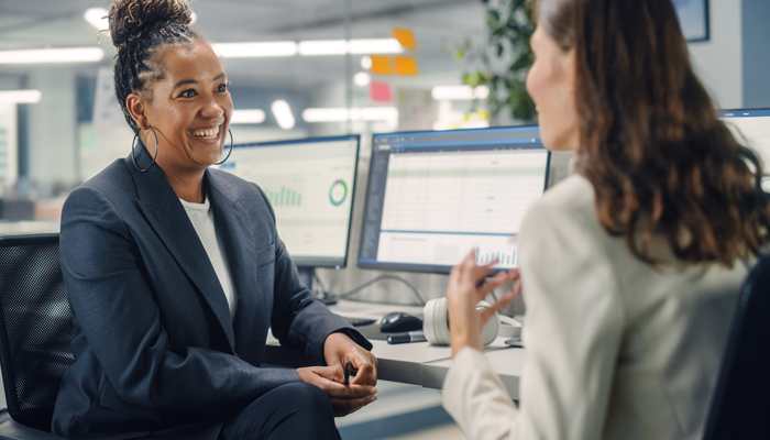 2 professional women, one black and one white smiling having a conversation sitting at a desk in an office