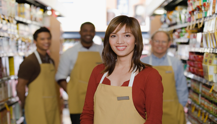 4 retail employees standing in a grocery isle. 3 mean blurred in back and one woman in focus smiling. all wearing retail aprons showcasing the canadian job market