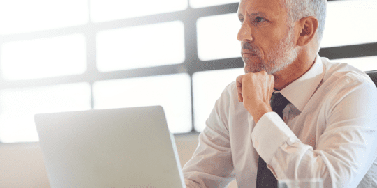 older male dressed in shirt and tie sitting at desk in front of laptop gazing ahead thinking about the labour market trends