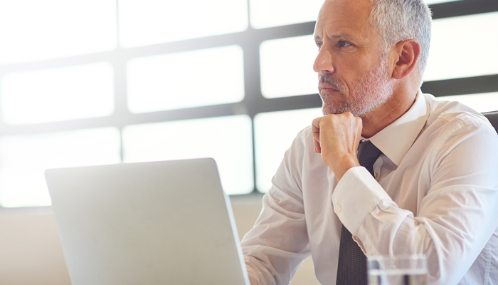 older male dressed in shirt and tie sitting at desk in front of laptop gazing ahead thinking about the labour market trends