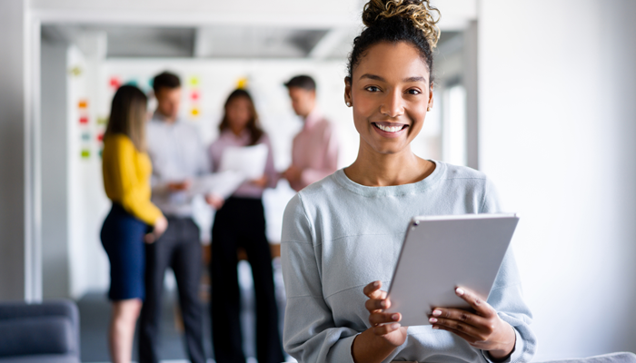 4 young professionals holding papers in blurred out background with one african american woman close up smiling holding a tablet