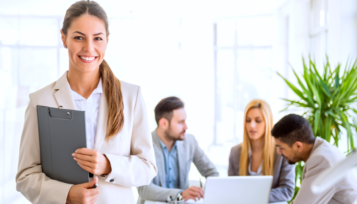 3 blurred out young professionals sitting around a desk and laptop and woman smiling holding clip board