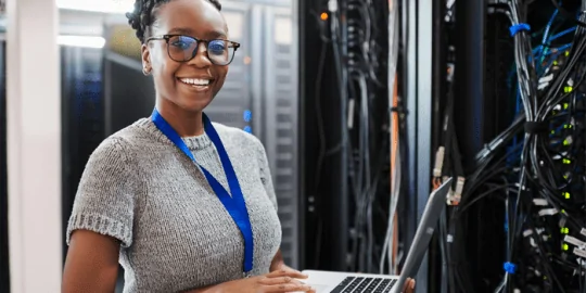young african american woman holding laptop smiling ahead with bundles wires against the wall