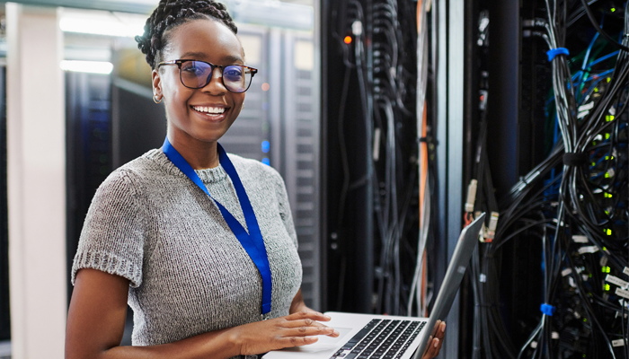 young african american woman holding laptop smiling ahead with bundles wires against the wall