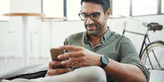 young man with glasses sitting on a sofa with bike in background holding mobile phone with slight smile