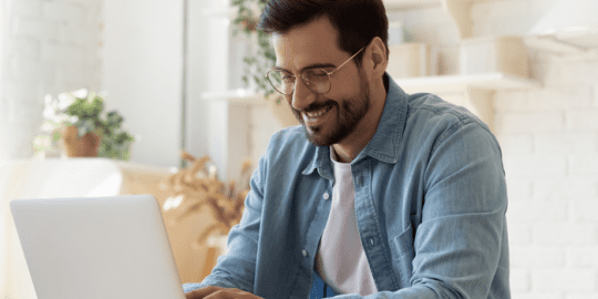 image of a young man with glasses dressed casually sitting desk working on laptop smiling