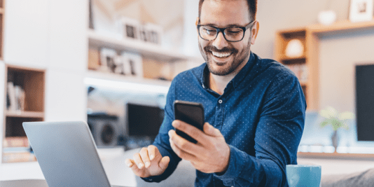 smiling man sitting in front of laptop looking at phone showcasing social media and the workplace
