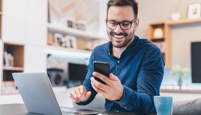 smiling man sitting in front of laptop looking at phone showcasing social media and the workplace