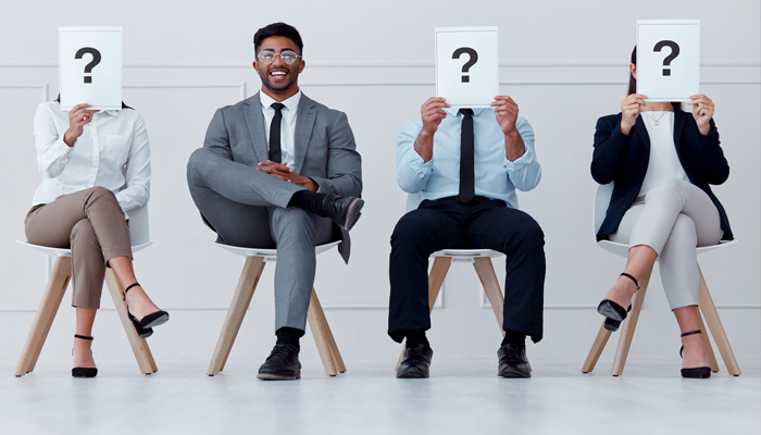 3 job candidates sitting in a row holding a question mark in front of their face and 1 man who is not and smiling as he completed a background check